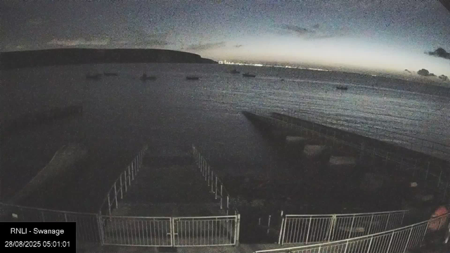 A view of a beach at dawn or dusk with calm water and a distant shoreline. The sky is partially illuminated, with darker clouds and a faint glow on the horizon. In the foreground, there is a fenced area with some structures on the beach. The scene appears peaceful with no people visible.