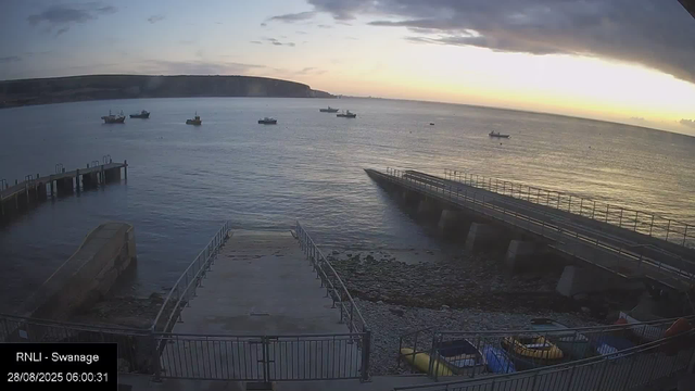 A sunrise view at a harbor with boats floating on the water. A pathway extends into the water on the right side, and there is a small pier on the left. In the foreground, there is a fenced area with colored kayaks or small boats. The sky is partly cloudy with the sun rising on the horizon.