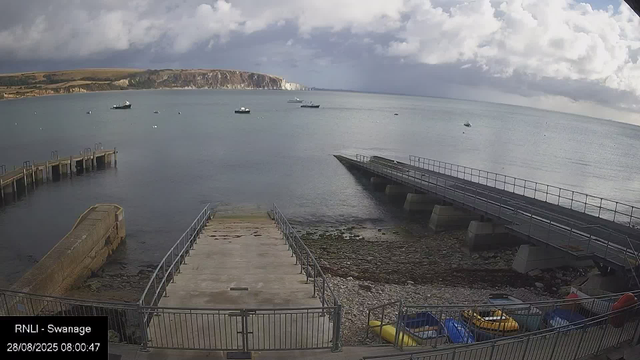 A tranquil harbor scene with a concrete pier extending into calm water. Several boats are anchored in the distance, and a rugged coastline with cliffs is visible under a partly cloudy sky. In the foreground, there are small parked boats and a metal railing surrounding the area. The timestamp indicates the image was captured on August 28, 2025, at 8:00 AM.
