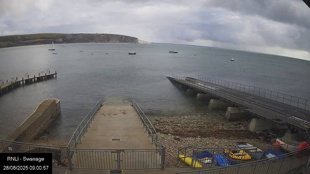 A view from a webcam overlooking a small harbor with boats on a cloudy day. There is a concrete pier extending into the water in the foreground, with a ramp leading down to the shore. Several colorful kayaks are stored in a fenced area near the bottom right corner. In the background, boats are floating on the calm sea, and a hillside with cliffs and some buildings stretches across the horizon under an overcast sky.