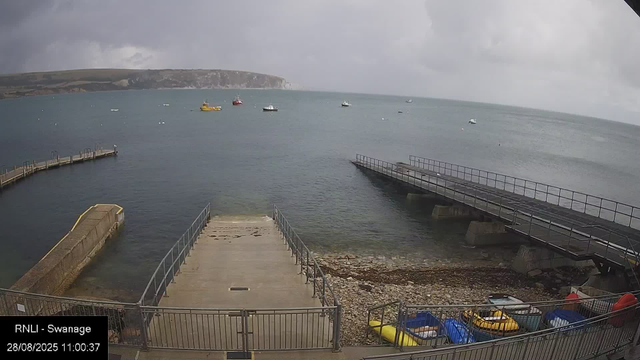 A view of a harbor with a concrete dock extending into the water, surrounded by metal railings. Several small boats are stored on the rocky shore near the dock. In the water, there are multiple boats and ships anchored in the distance. The sky is cloudy with dark clouds, suggesting an overcast or stormy weather. A large landmass or cliff is visible across the water in the background. The timestamp indicates the photo was taken on August 28, 2025, at 11:00 AM, and the location is identified as Swanage by RNLI.