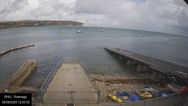 A view of a harbor with a concrete pier extending into the water from the bottom center, flanked by metal railings. To the right, there's a small boat on the rocky shore. Several boats are anchored in the calm water, and a yellow boat is visible among them. In the background, there's a distant coastline with cliffs and hills under a cloudy sky.