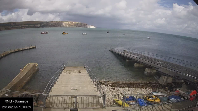 A view of a small harbor with calm water, several boats anchored in the distance, and a coastline with cliffs in the background. There are two concrete piers extending into the water, one on the left with a person standing at the end, and a longer one on the right. In the foreground, there are colorful small boats, kayaks, and paddleboards stored behind a metal fence. The sky is partly cloudy, with patches of sunlight breaking through. The timestamp indicates the time as 13:00:39 on August 28, 2025, and it is labeled "RNLI - Swannage."