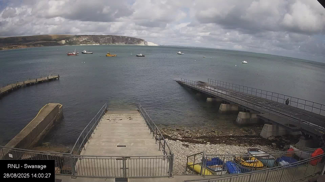 A view of a harbor with a concrete pier extending into the water, flanked by metal railings. Several small boats are docked or floating nearby, with some colorful kayaks and paddle boats visible in a fenced area at the bottom right. In the distance, multiple boats are on the water, and a large cliff with white chalky cliffs and green hills rises on the horizon under a partly cloudy sky.