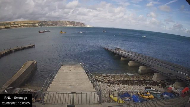 A view of a harbor with a concrete pier and a small dock extending into the water. Several boats and ships are floating on the water in the distance, and rolling hills with cliffs are visible on the horizon under a partly cloudy sky. There are some small colorful boats or kayaks stored in a fenced area at the bottom of the image.