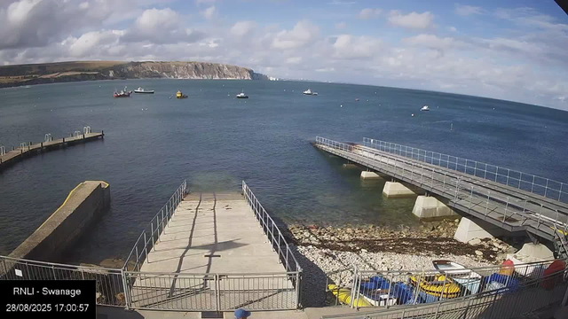 A view of a harbor with a concrete pier extending into calm water. Several boats are anchored in the water, with a rugged coastline and cliffs visible in the background. The sky is partly cloudy with patches of blue. At the bottom of the image, there is a fenced area with colorful paddle boats and other watercraft. The timestamp shows the date as August 28, 2025, at 17:00:57.