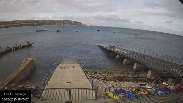 A seaside scene showing a small harbor with a concrete dock extending into the water, a boat ramp leading into the water, and a collection of colorful paddle boats and kayaks stored behind a fence in the foreground. Several boats are anchored in the calm sea, and a cliff with grassy slopes is visible in the distance under partly cloudy skies.
