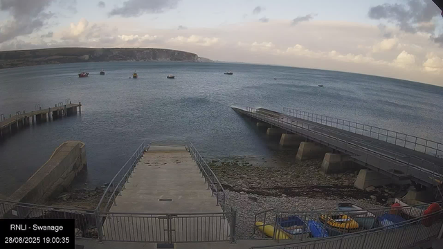 A view of a harbor with a small concrete and metal pier extending into calm water. Several boats are floating in the distance on the water, with a large cliff or headland visible on the horizon. The sky is partly cloudy, with patches of sunlight breaking through clouds. In the foreground, there are parked cars, including yellow, blue, and red vehicles, next to a fenced area. The image is timestamped "28/08/2025 19:00:35" and labeled "RNLI - Swansea."