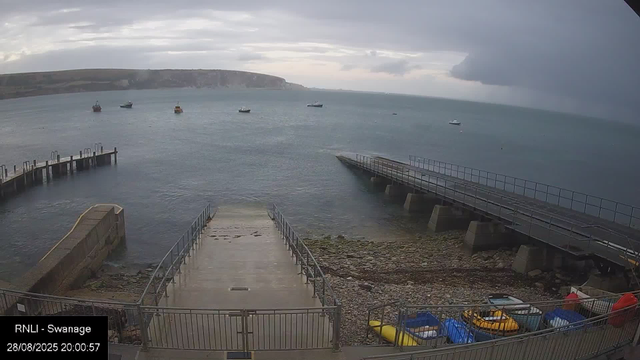 A view of a harbor with a concrete dock extending into the water, flanked by metal railings. Several small boats are floating in the water, and a rocky shoreline is visible beneath the dock. In the background, there are cliffs and a cloudy sky, suggesting an overcast evening. At the bottom left corner, there is a timestamp and location label reading "RNLI - Swanage, 28/08/2025 20:00:57."