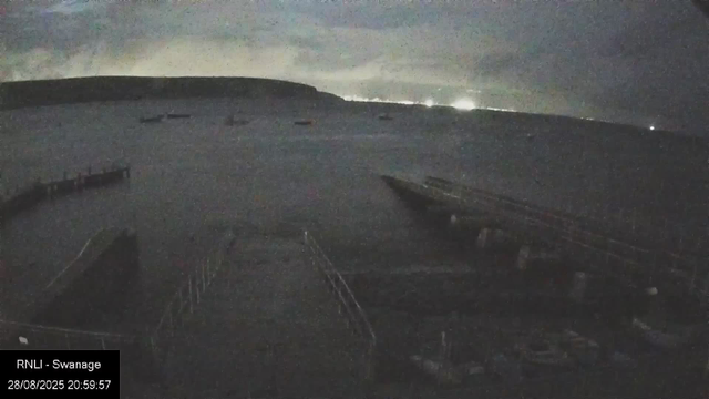 A dark, nighttime view from a webcam showing a mountain landscape with a few small buildings or structures on the slope. The sky is cloudy with limited visibility of stars or celestial objects. There is a faint glow of distant lights or lightning on the horizon. The bottom left corner has a timestamp and location information: "RNLI - Swanage, 28/08/2025 20:59:57".