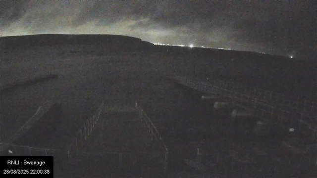 A dark nighttime sky with faint clouds above a large, dark aircraft viewed from the front cockpit. The horizon shows a few distant lights, and the bottom left corner has a timestamp and location text.