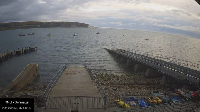 A view of a harbor with a concrete pier extending into the water, a few small boats floating in the bay, and a rocky shoreline. In the background, there are cliffs and a cloudy sky with patches of sunlight breaking through. At the bottom left corner, there is a timestamp and location label indicating the photo was taken at Swanage on August 29, 2025, at 07:00:09.
