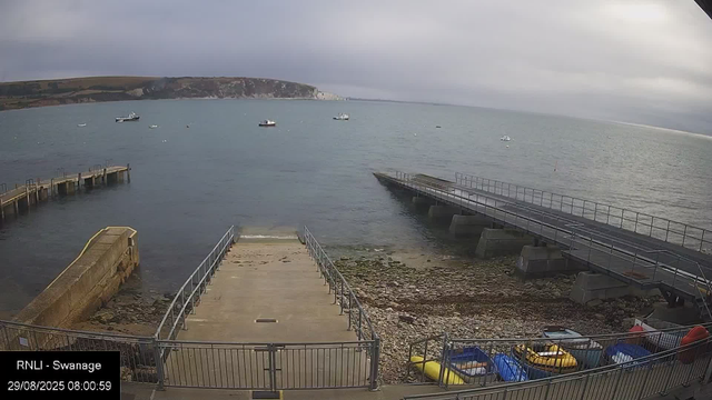 A view of a harbor with a small dock extending into the water, surrounded by a railing. To the right, there is a sloped boat ramp with a metal railing. The shoreline has rocks and pebbles, with several colorful kayaks or small boats stored below. The water is calm with a few boats anchored in the distance. In the background, there are cliffs or headlands under a cloudy sky. The timestamp in the bottom left corner indicates the date and time as 29/08/2023 at 08:00:59.