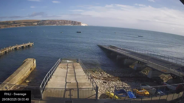 A view of a seaside harbor with a concrete pier extending into the calm ocean water. Several boats are floating in the distance. To the right, a metal walkway or slide descends into the water. In the foreground, there's a fenced area with parked cars on a rocky shoreline. The background shows a cliff overlooking the sea under a partly cloudy sky, with gentle waves lapping against the shore.