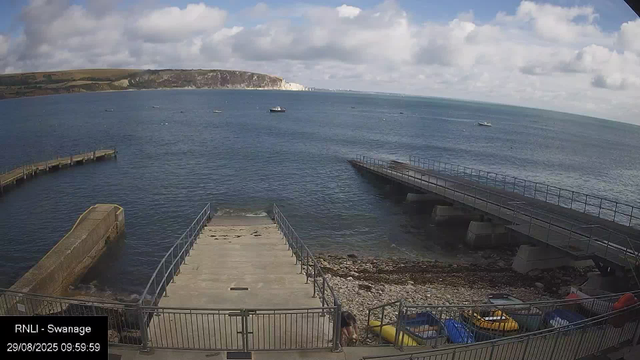A view of a seaside area with a small pier extending into the water. There are boats floating in the bay and a distant shoreline with cliffs and hills. The sky is partly cloudy with patches of blue.
