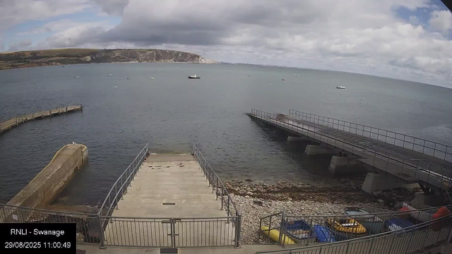 A view of a seaside dock area with railings and a concrete platform leading into the water. Several small boats and kayaks are stored along the edge, and a few larger boats are floating in the water. In the background, there are cliffs and a cloudy sky above the sea.