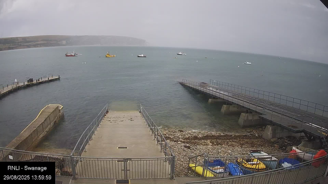 A view of a harbor with two concrete piers extending into the water, one on the left and one on the right. The water is calm with a few small boats floating on it. In the foreground, there are colorful kayaks and small boats stored behind a metal fence, and a rocky shoreline is visible below the piers. The sky is overcast, and a distant landmass can be seen across the water.