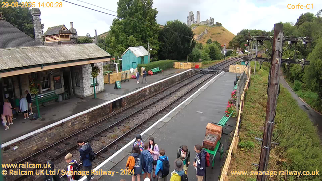 A small train station platform with a stone building on the left, several groups of people standing and chatting, some with backpacks. There are flower pots and benches along the platform, as well as a small green shed. A line of train tracks runs through the scene, and an overhead utility pole with wires is visible on the right. In the background, there are green trees, a hillside, and ruins of an old castle on top of a hill. The sky is overcast.