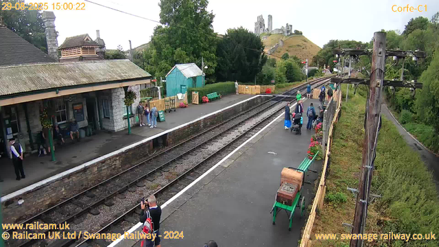 A small train station platform with a few people waiting, some standing near the entrance building, and others further down the platform. There are benches, flower pots, and a small turquoise shed in the background. A person is taking a photo or video with a camera, and some passengers are pushing strollers or carrying luggage. The railway tracks run through the center of the image, with a backdrop of greenery, trees, and a hill with ruins or a castle on top. Utility poles with electrical wires run along the right side of the platform. The sky appears partly cloudy. The image has a timestamp of August 29, 2025, at 15:00.