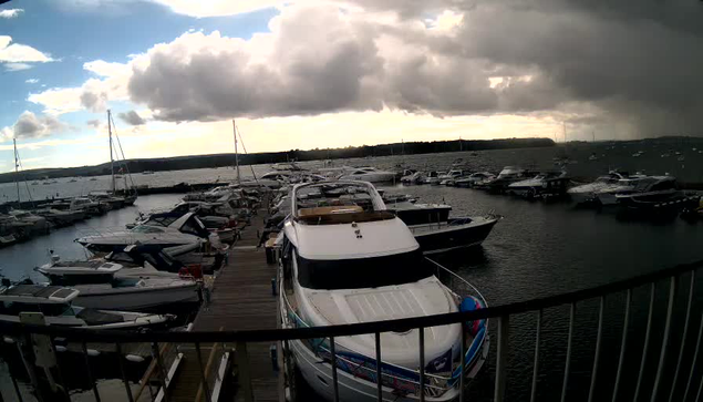 A marina with numerous boats docked along wooden piers. The boats vary in size, with some sailing yachts and smaller motorboats. The sky is partly cloudy with large, dark clouds, and the water appears calm. A railing is visible in the foreground.