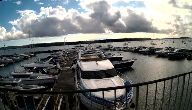 A marina with multiple boats docked along a pier extends into a body of water. The boats vary in size and style, some with covered cabins. The sky overhead is partly cloudy with large, dark clouds, and there is a distant shoreline visible on the horizon. A railing is visible in the foreground, indicating the photo was taken from an elevated position.