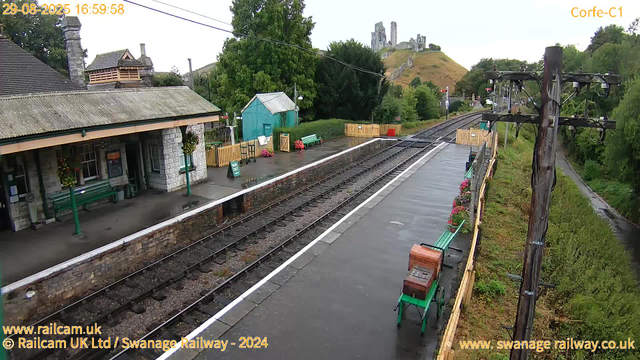 A quaint railway station with a single platform, surrounded by lush green trees and rolling hills. The station building is made of stone with a tiled roof, and has green benches and flower baskets hanging outside. There is a small teal-colored shed on the platform, with flower arrangements and a yellow picket fence. Overhead, there are railway power lines and a tall wooden utility pole. In the distance, atop a hill, are the ruins of an old castle. The weather appears to be overcast, and the platform looks wet, indicating recent rain.