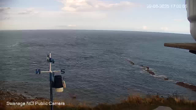 A view of the ocean from a webcam shows a vast body of water extending to the horizon under a partly cloudy sky. In the foreground, there is a weather vane and an anemometer mounted on a pole at the edge of a coastal cliff. A small boat or vessel is seen traveling across the water, creating a trail behind it. The image also includes a timestamp in the top right corner, indicating the date and time as 29-08-2025 17:00:06. The bottom left corner has a label reading "Swanage NCI Public Camera."