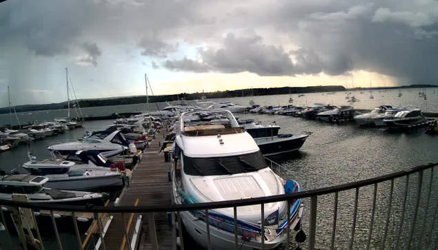 A large white yacht is docked at the end of a wooden pier, surrounded by smaller boats and sailboats on a calm body of water. Dark clouds loom overhead with a hint of sunlight breaking through in the distance. The scene appears to be in a marina or harbor.