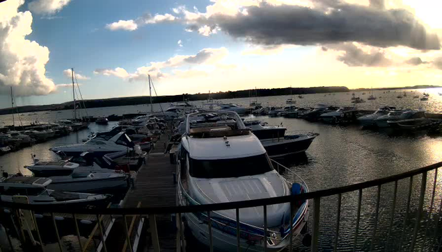 A harbor filled with numerous boats docked at a wooden pier. The sky is partly cloudy with the sun setting on the horizon, casting a warm glow over the water. A railing is visible in the foreground.