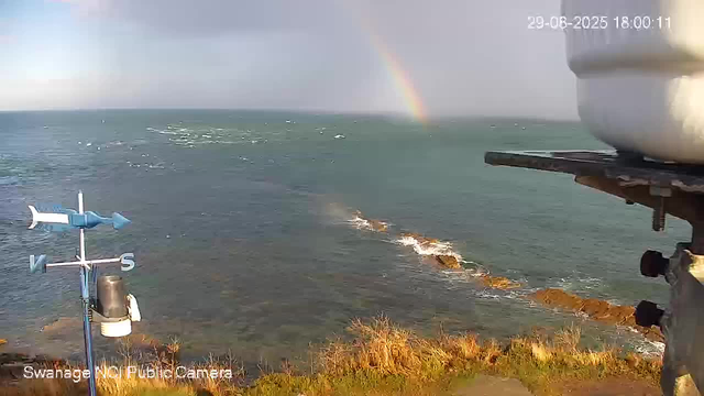 A view from a webcam showing a body of water with small waves and rocks along the shoreline. In the distance, a rainbow arcs across a sky with dark clouds. There is a weather vane with directional markers (N, E, S, W) and an anemometer situated near the edge of the scene.