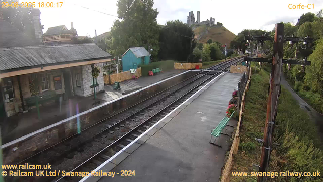 A quaint train station platform on a cloudy day, with a small stone building on the left, decorated with hanging flower baskets. There are a few benches along the edge of the platform, some with potted flowers nearby. Parallel railway tracks run through the center, curving into the distance towards a hillside with castle ruins or historic structures atop it. To the right, there's a tall wooden utility pole with electrical wires and hanging insulators, and a narrow footpath runs alongside it. The station appears quiet and well-maintained, with a mix of lush green trees and hillside scenery in the background.
