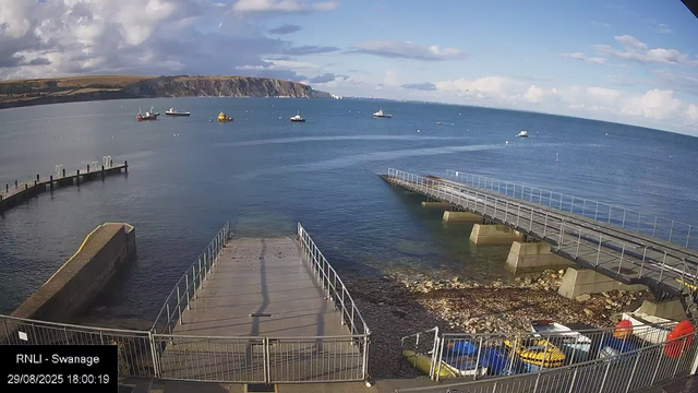 A view of a coastal scene with calm blue water and several boats anchored offshore. In the foreground, there is a small pier and a concrete slipway, with a few colorful boats parked underneath a metal fence. The sky is partly cloudy with patches of blue and distant cliffs along the horizon.