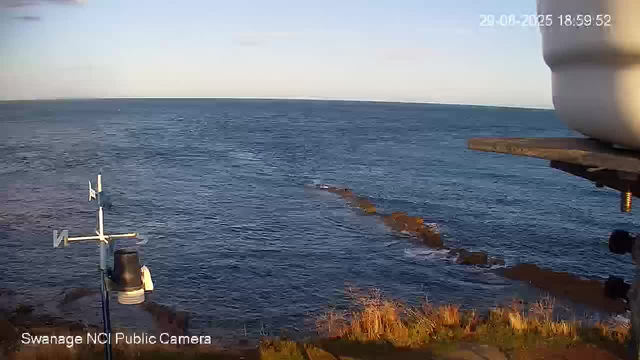 A view of the ocean with rocky formations extending into the water. The scene is bathed in warm, golden light, suggesting it is late afternoon or early evening. In the foreground, there is a weather station with an anemometer and other instruments mounted on a pole. The timestamp on the image shows the date as January 20, 2025, at 6:39 PM. The camera is labeled "Swanage NCI Public Camera."