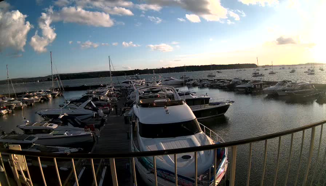 A marina with multiple boats and yachts docked, seen from a wooden pier. The sky is partly cloudy with the sun setting on the horizon, casting a warm glow over the scene. The water is calm, reflecting the boats and sky. A curved metal railing is in the foreground.