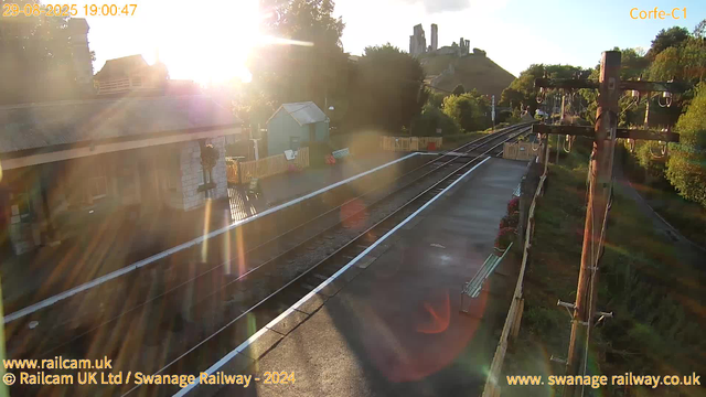 A deserted railway station platform bathed in the warm glow of late afternoon sunlight. The tracks run diagonally from the foreground to the background, flanked by a weathered utility pole with hanging wires on the right side. On the left, there are a few benches and small shelters, with a yellow safety fence separating the platform from surrounding greenery. In the distance, a castle-like ruin perches atop a hill with trees colored by the autumn season, under a clear sky. The sun’s rays create lens flare effects across the image.