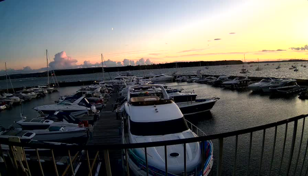 A marina filled with numerous boats docked along wooden piers during sunset. The sky is painted with soft hues of orange, pink, and blue, with some clouds and a faint crescent moon visible. The water is calm, reflecting the boats and the colorful sky. In the foreground, a railing runs along the edge of the dock.
