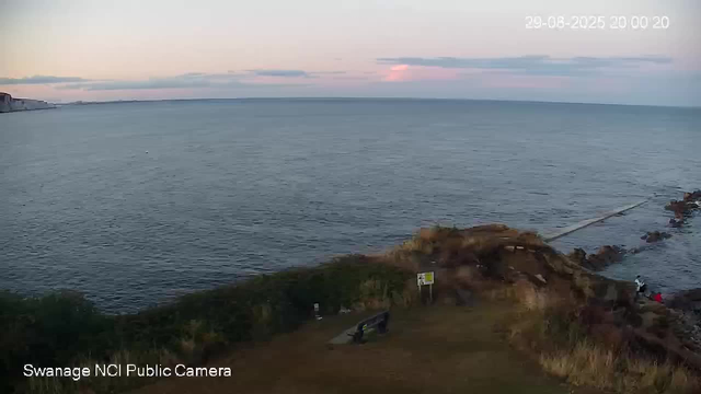 A view of the coastline during sunset, with calm ocean waters stretching to the horizon. In the foreground, there is a grassy area with a bench and a small sign. Rocks and a narrow pathway lead toward the water on the right side, where a person is sitting near the shoreline. The sky is partly cloudy with hues of pink and blue. The timestamp in the top right corner shows the date and time as 29-08-2026 at 20:00:20.