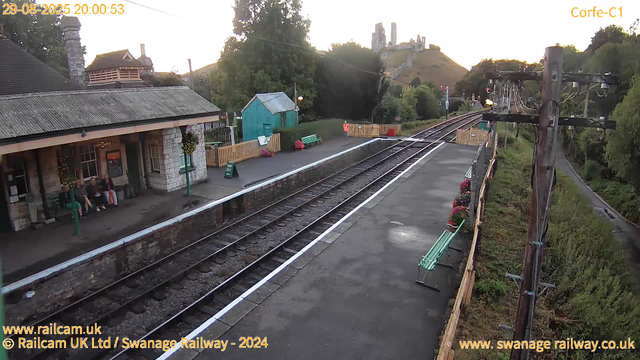 A small train station platform with a building on the left, where several people are sitting. There is a blue shed, flower pots, and a green bench on the platform. A person in a high-visibility vest stands near the edge of the platform. Overhead are electric wires supported by tall wooden poles. In the background, a hill with a castle ruin or historic structure is visible. The time and date are displayed in the top left corner: 29-08-2025 20:00:53.