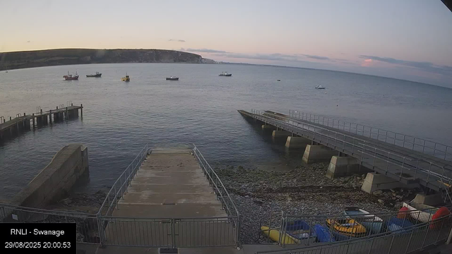 A view from a webcam showing a calm seaside scene at dusk. There are several boats floating on the water, with a larger landmass or cliff in the distance. In the foreground, there are two piers extending into the water, with a small concrete area and some colorful kayaks or watercraft stored along a fence on the right side. The sky is partly cloudy with soft pastel colors. The bottom left corner displays the text "RNLI - Swanage" along with the date and time: 29/08/2025 20:00:53.