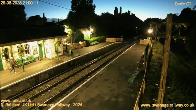 An evening at a small railway station on August 29, 2025, at 9:00 PM. The platform is illuminated by a few warm lights, with a stone building on the left side, featuring green benches, potted plants, and lantern-style lamps. A person is walking across the platform near the building. The tracks run down the center of the image, with a curved section heading into darkness. On the right side, wooden utility poles support overhead wires, and there are flower baskets hanging along a fence. The background shows a dark sky transitioning to twilight, with silhouettes of trees and some structures on a hill in the distance.