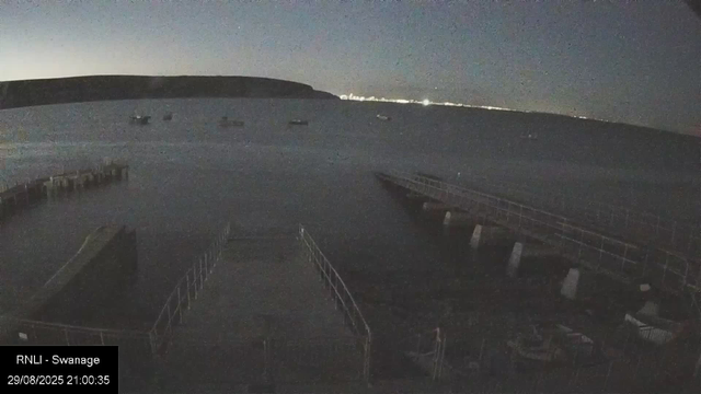 A nighttime view of a harbor with several small boats floating on calm water. There are empty walkways and railings along the edge of the harbor. In the background, a dark hill or shoreline is visible, with a few distant lights along the horizon. The sky is dark, and the overall scene is dimly lit. A label in the bottom left corner reads "RNLI - Swannage" with a date and time of 29/08/2025 21:00:35.