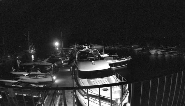 A marina at night with several boats and yachts docked. The scene is illuminated by a few bright lights, creating reflections on the water. The boats vary in size and are positioned close together along the dock.