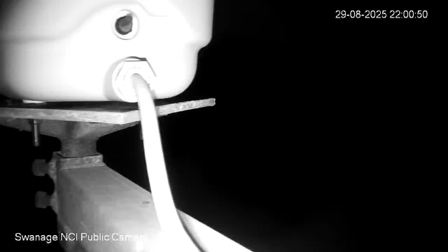 A black and white image from a webcam showing a close-up view of a mechanical or industrial device. There is a large, round, white component on the left side with a small dial or sensor near the center. A tube or pipe extends downward from this component. The device appears to be mounted on a metal platform or arm, with visible bolts and brackets. The background is dark, and a timestamp in the upper right corner reads "29-08-2025 22:00:50." The bottom left corner has the text "Swanage NCI Public Camera."