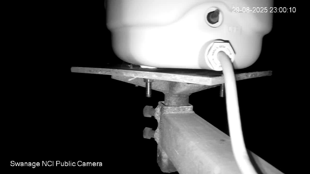 A close-up black-and-white image captured by a webcam shows a white device, possibly a sensor or camera, mounted on a metal structure with bolts. The background is dark, and there is a timestamp in the upper right corner indicating the date and time as 29-08-2025 23:00:10. The bottom left corner has text reading "Swanage NCI Public Camera."