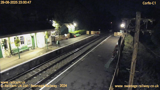 A quiet railway station platform at night, illuminated by bright streetlights. To the left, there is a white building with green doors, windows, and benches outside, along with potted plants and a small green sign. In the center, railway tracks run parallel to the platform, which has a safety line along the edge. On the right, there is a tall utility pole with wires, and beyond it, darkness. The top left corner displays the date and time as "29-08-2025 23:00:47".