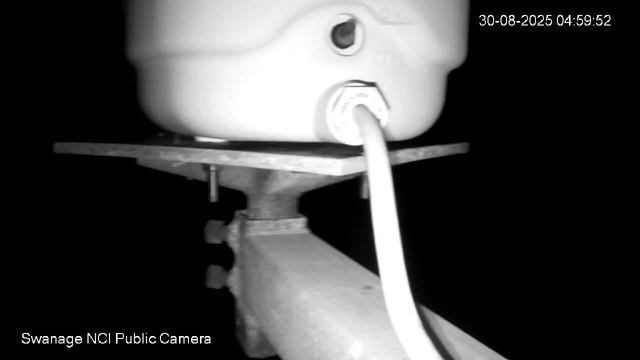 A black and white image from a webcam showing a close-up of a mounted device connected by a cable. The device appears to be attached to a wooden or metal structure. The background is dark, and the timestamp in the upper right corner shows the date "30-08-2025" and the time "04:59:52." The bottom left corner has the text "Swanage NCI Public Camera."