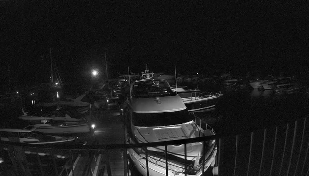 A black-and-white image of a marina at night shows several yachts and boats docked closely together. The scene is dark, with some boats faintly illuminated by distant light sources. A railing is visible in the foreground, framing the view of the boats on the water.