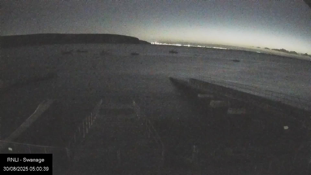 A nighttime view from a drone shows a wide, open landscape with a gently rolling terrain. In the distance, some faint lights are visible on the horizon, while the sky is dark with a subtle gradient near the horizon, suggesting dawn or dusk. The image is grainy and mostly dark, with the silhouette of the drone's landing gear visible at the bottom of the frame. The timestamp indicates it was captured early morning on August 30, 2025.