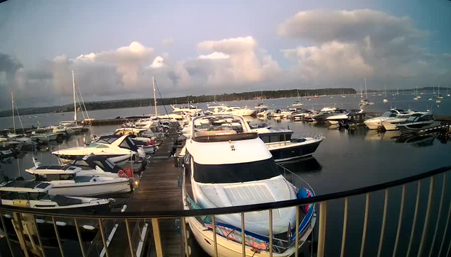 A marina filled with numerous white and dark-colored yachts and boats docked alongside wooden piers. The water is calm, reflecting the boats, and the sky above is partly cloudy with large, fluffy clouds. The view is from a vantage point behind a metal railing.