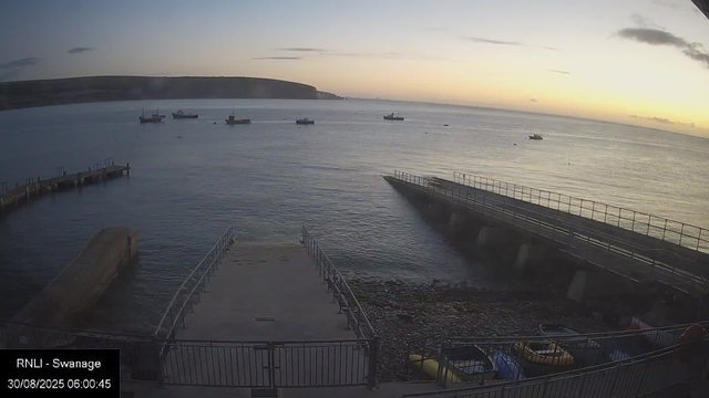 A view of a calm sea at dawn with a few boats floating on the water. In the foreground, there is a concrete ramp leading into the water, surrounded by a metal railing. To the right, a larger ramp slopes into the sea, also with a railing, and some boats are visible below on the rocky shore. The sky is partly cloudy with the sun just beginning to rise, casting a soft glow over the scene. In the top left corner, there is a distant shoreline with cliffs. In the bottom corner, a timestamp and location are displayed: "RNLI - Swanage, 30/08/2025 06:00:45."